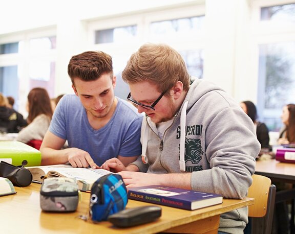 Zwei Auszubildende sitzen in der Schule an einem Tisch und schauen in ein Buch 