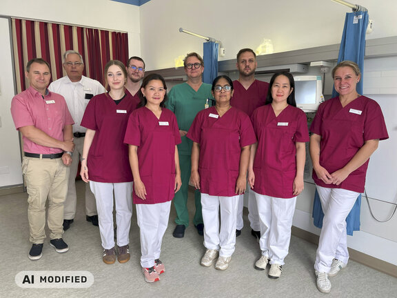 Das Bild zeigt eine Gruppe von Personen in einem Patientenzimmer. Zu sehen sind Pflegefachkräfte in dunkelroten Kasacks sowie Mitarbeitende des Qualitätsmanagements und der Pflegedirektion sowie der Leitende Kardiologe in einem grünen Kasack.