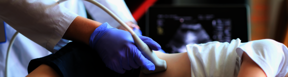 Man sieht die Leiste einer Patientin und eine Arzt, der mit einem Ultraschallgerät den Seitlichen Bauchbereich abgeht. Es sind keine Gesichter zu sehen und im Hintergrund sieht man einen Bildschirm, auf dem man die Ultraschallergebnisse sieht. 