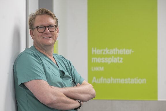 Dr. med. Frithjof Schlegel, Leiter der Kardiologie, steht mit verschränkten Armen und in die Kamera blickend vor dem Schild, das auf den Herzkathetermessplatz im St.-Clemens-Hospital verweist.