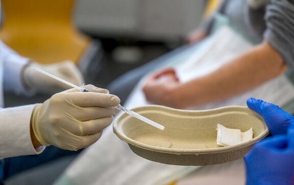 Neurologischer Arzt hält eine Nierenschale und eine Spritze in der Hand. Im Hintergrund sieht man den Arm eines Patienten. 