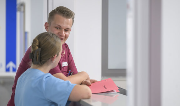 Pflegefachkräfte stehen an einem Stationsempfang und reden miteinander. 