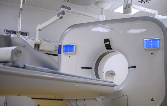 Bild des MRT von Vorne ohne Patienten. Die Liege ist ausgefahren und die Bildschirme des MRT Gerätes leuchten Blau. 
