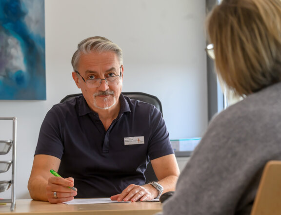 Neurologischer Facharzt führt ein Gespräch mit einer Patientin. Dabei schaut er in die Kamera. 